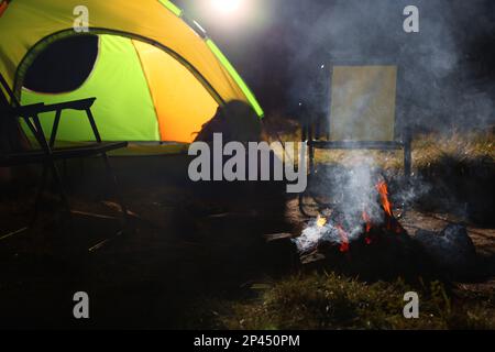 Falò per fumatori e sedie pieghevoli vicino alla tenda da campeggio all'aperto di sera Foto Stock