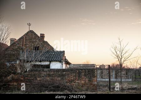 Foto di una fattoria abbandonata in Vojvodina, in Serbia, con la facciata della sua casa principale gravemente danneggiata e decadente. Foto Stock