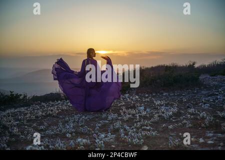 Tramonto viola abito donna montagne. Ascesa del mistico. tramonto sulle nuvole con una ragazza in un lungo vestito viola. Nel prato c'è un prato Foto Stock