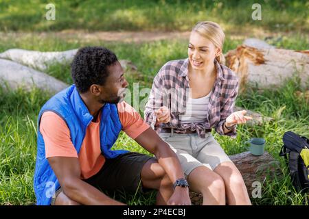 Felice, coppia diversa seduta sul log in foresta soleggiata, parlando Foto Stock