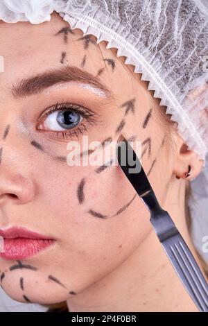 Primo piano del bisturi vicino alla faccia delle giovani donne. Concetto di funzionamento di bellezza. Ritratto di donna con linee chirurgiche sulla pelle. Foto Stock