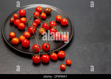 Rametto di pomodori ciliegini rossi su un piatto nero. Spazio di copia. Giacitura piatta. Sfondo nero. Foto Stock