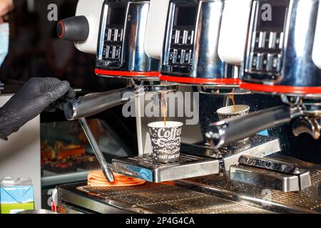 La mano di un barista in un guanto in lattice nero versa il caffè espresso caldo in una tazza di carta da una macchina da caffè. Foto Stock