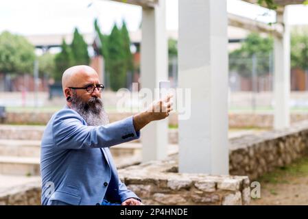 uomo di mezza età, calvo, bearded seduto a prendere selfie nel parco Foto Stock