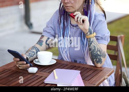 Giovane donna con tatuaggi sulle braccia che testano in smartphone e fumando vape pen mentre si siede al tavolo in un bar all'aperto e si serve un caffè Foto Stock