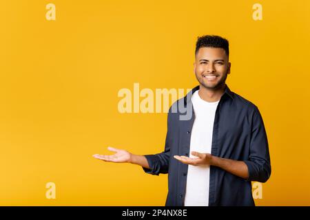 Felice eccitato giovane uomo indiano guardando la macchina fotografica che punta da parte con entrambe le mani, gesturing allo spazio della copia che fa pubblicità alla nuova promozione, presentando l'offerta di vendita in piedi isolato su sfondo giallo. Foto Stock