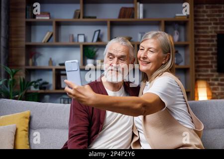 Coppia matura e felice che si ingoola sulla fotocamera dello smartphone. Marito e moglie dai capelli grigi stanno parlando facendo selfie divertenti. Videochiamata sullo smartphone Foto Stock