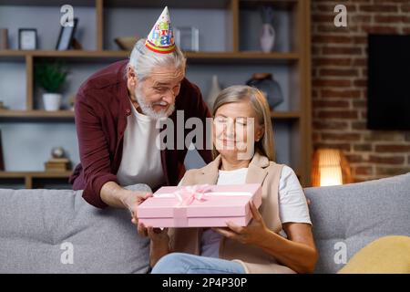 Premuroso vecchio marito dando romantico regalo di compleanno a moglie matura eccitato, amore uomo più anziano sorpresa felice donna di mezza età, scatola rosa aperta, bacio Foto Stock