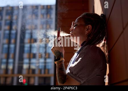 Giovane donna con vape pen in mano in piedi dal muro di costruzione e rilasciando nube di vapore dalla sua bocca mentre fuma Foto Stock