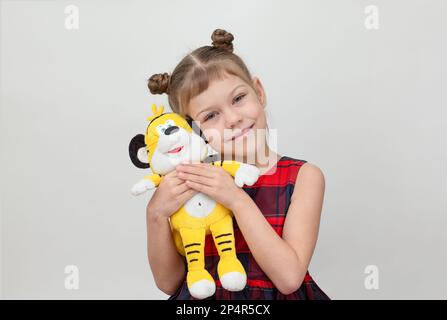 Bambino felice e sorridente che abbraccia il giocattolo della tigre su sfondo bianco bambina caucasica di 6 7 anni in vestito rosso plaid guardando la macchina fotografica Foto Stock