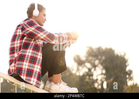 Giovane donna a capelli corti in cuffie seduta su ringhiera all'aperto e utilizzando il suo telefono. Foto Stock