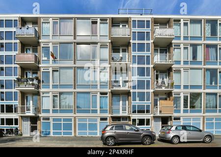 Amsterdam, Paesi Bassi - 10 aprile 2021: Due auto parcheggiate di fronte ad un edificio di appartamenti a più piani con molte finestre e balle laterali Foto Stock