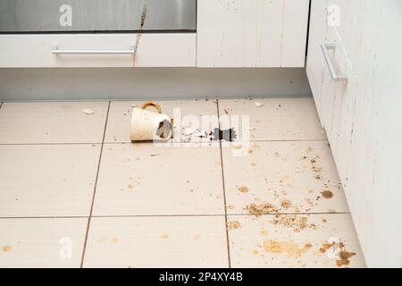 Tazza da tè rotta sul pavimento della cucina, tazza da caffè schiacciata e caffè macinato su tutte le piastrelle. Foto Stock