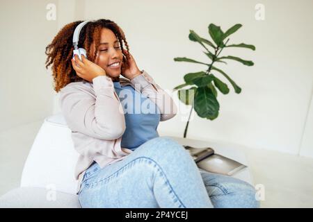 Corpo positivo curvy afro donna americana in cuffie su un divano leggero a casa. Giovane donna scura con pelle in abiti casual chiuse gli occhi e gode t Foto Stock