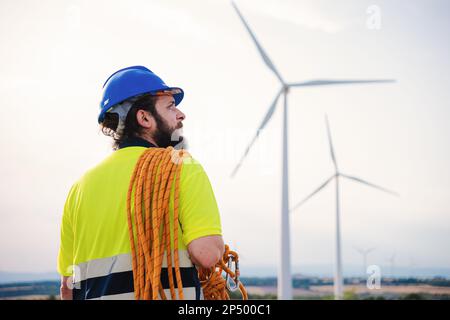 Vista posteriore del tecnico di manutenzione delle turbine a vento in piedi indossando un elmetto e un giubbotto di sicurezza presso una fattoria eolica con attrezzatura per arrampicata. Foto di alta qualità Foto Stock