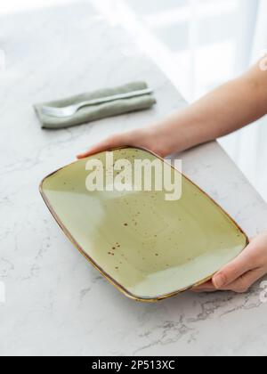 Mani che trattengono il piatto rettangolare verde vuoto con forchetta su tavolo in marmo bianco davanti alla finestra. Vista dall'alto Foto Stock