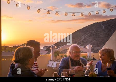 Un gruppo felice di persone anziane si godranno il tempo della cena con il tramonto e la città sullo sfondo. Anziani maturi stile di vita coppie uomini e donne che mangiano e bevono Foto Stock