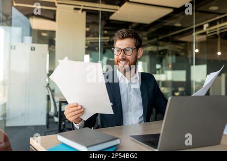 Felice imprenditore maschio che legge i documenti di affari e sorridente, controllando i tassi di crescita dell'azienda, seduto alla scrivania in ufficio Foto Stock