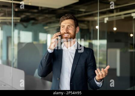 Uomo d'affari caucasico di successo che parla sullo smartphone, in piedi in ufficio all'interno, avendo conversazione telefonica Foto Stock
