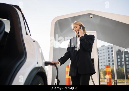 Felice donna di affari caucasica con capelli biondi utilizzando uno smartphone moderno per la conversazione, mentre in attesa di auto elettrica a caricare all'aperto EV stazione. Concetto di ecologia e di trasporto. Foto Stock