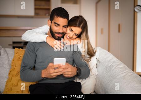 Sorridente donna europea millenaria abbraccia arabo marito gioca a gioco su smartphone, legge messaggio Foto Stock