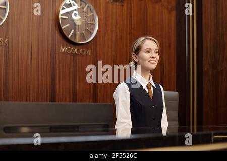 Giovane receptionist femminile di successo in piedi uniforme presso il banco check-in e registrazione, mentre in attesa di nuovi ospiti o turisti Foto Stock