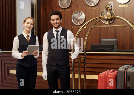 Giovane e felice, elegante receptionist e fattorino in piedi nella lounge di un hotel di lusso e guardando la macchina fotografica mentre aspettate nuovi ospiti Foto Stock