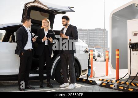 Lavoratori multirazziali in tute intelligenti con dispositivi moderni in piedi vicino all'e-car. Il compagno di squadra arabo attende che l'auto ecologica venga ricaricata con energia presso un luogo di ricarica appropriato. Foto Stock