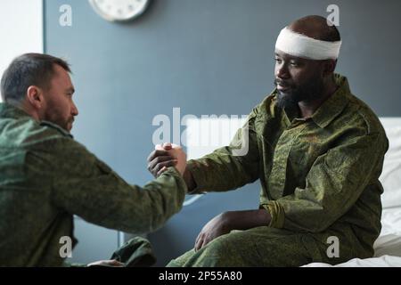 Giovane soldato che scuote la mano dell'amico afro-americano con la testa ferita mentre si siede di fronte a lui durante la visita in reparto ospedaliero Foto Stock