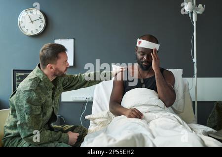 Giovane uomo in uniforme militare che sostiene il suo amico ferito seduto a letto e tenendo la mano sulla testa ferita avvolta con bendaggio Foto Stock