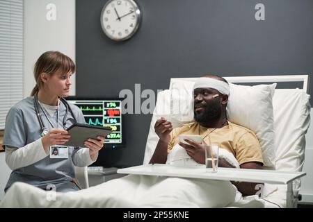 Uomo afroamericano ferito con testa bandita che mangia la colazione a letto e che parla con l'infermiera che inserisce i dati in tablet nel reparto dell'ospedale Foto Stock