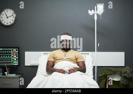 Giovane soldato afroamericano con ossimetro sulla punta delle dita e bendatura sulla testa seduta a letto nel reparto dell'ospedale e guardando la macchina fotografica Foto Stock