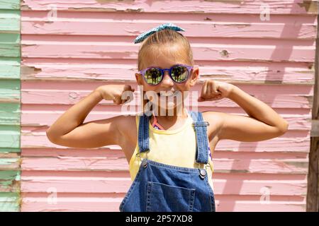 Ritratto di felice ragazza biraciale sorridente e mostrando i muscoli sopra la parete rosa Foto Stock