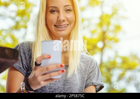 Bella ragazza bionda sorridente in una foresta all'aperto o in un parco pubblico in una giornata di sole. Ha bretelle molto visibili che la rendono ancora più bella. Ha un SM Foto Stock