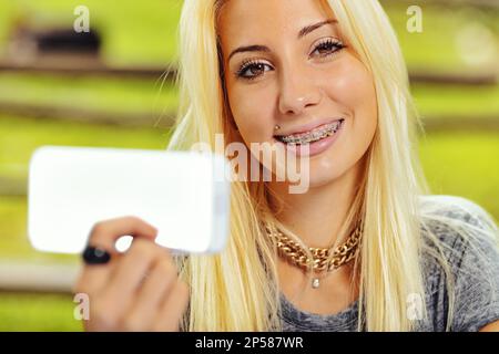 Sorridente ragazza con dispositivo mostra uno schermo vuoto per copiare lo spazio dello smartphone fuori messa a fuoco in primo piano. È bionda e bella, molto giovane e all'aperto Foto Stock