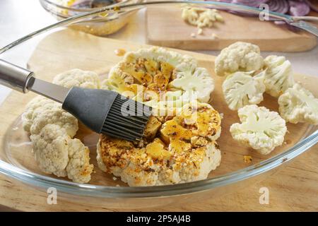 Le fette di cavolfiore crudo in una casseruola di vetro sono ricoperte con una miscela di spezie, erbe e olio d'oliva da una spazzola di silicone, preparazione per la cottura in forno Foto Stock