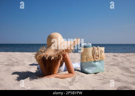 Donna con borsa da spiaggia e cappello di paglia sdraiato sulla sabbia vicino al mare Foto Stock