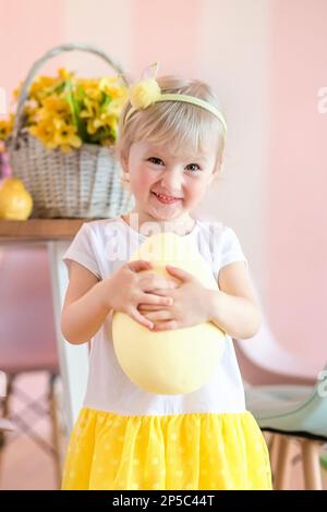 Una ragazza piccola divertente tiene un uovo giallo grande in una cucina decorata per la festa di Pasqua. La tradizione di trovare le uova per una festa religiosa. Foto Stock