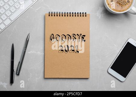 Notebook con frase Goodbye 2021, smartphone, tastiera, penne e tazza di caffè su tavolo in marmo grigio, piatto Foto Stock