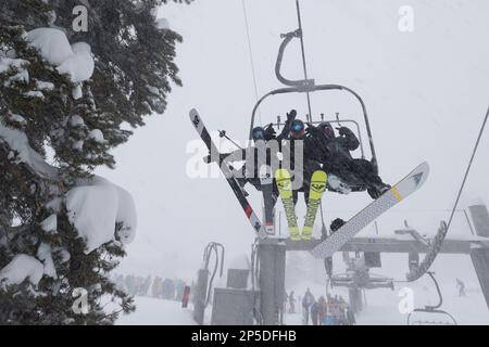 Con una fila di persone che aspettano dietro, due sciatori e uno snowboarder cavalcano uno skilift durante una buzzard invernale presso la stazione sciistica di Mammoth Mountain. Foto Stock