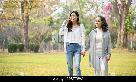 Bella giovane nipote asiatica tiene le mani con la nonna mentre passeggi nel bellissimo parco verde insieme. Foto Stock