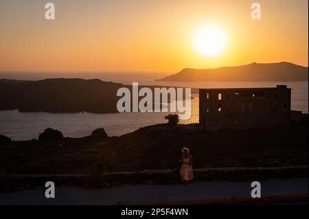 Vista al tramonto della città di Fira sull'isola greca di Santorini. Una donna siede di fronte ad un edificio vuoto della vecchia miniera di Fira, che si trova nella fronte Foto Stock