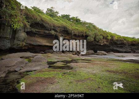 Foto del complesso del tempio pura Tanah Lot, nelle pietre ricoperte di muschio in primo piano e nell'acqua, sullo sfondo le scogliere verdi. Foto Stock