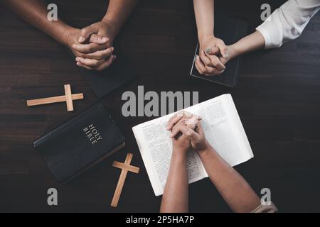 Piccoli gruppi di cristiani tengono le mani e pregano insieme intorno ad un tavolo di legno con pagine bibliche e croci. Gruppo di studio biblico imparare conce religiose Foto Stock