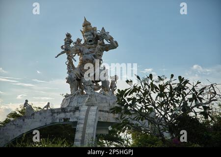 Foto di una statua di scimmia indù presso il complesso del tempio pura Luhur a Uluwatu a Bali, in primo piano in modo diffuso una pianta, sullo sfondo il cielo blu. Foto Stock