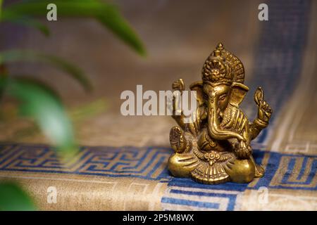 Statua in bronzo di ganesha su un tappeto d'oro con una pianta verde in primo piano Foto Stock