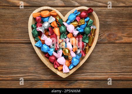 Piatto a forma di cuore con caramelle in involucri colorati su tavolo in legno, vista dall'alto Foto Stock