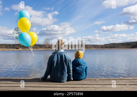 un ragazzino e una donna irriconoscibili si siedono con le spalle sulla riva di un lago con palloncini blu e gialli. Famiglia, rifugiati, unità, sostegno, patri Foto Stock