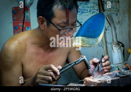 Un gioielliere e oreficeria lavora su un pezzo d'oro con una torcia a soffio e pinzette all'interno del mercato centrale di Phnom Penh, Cambogia. Foto Stock