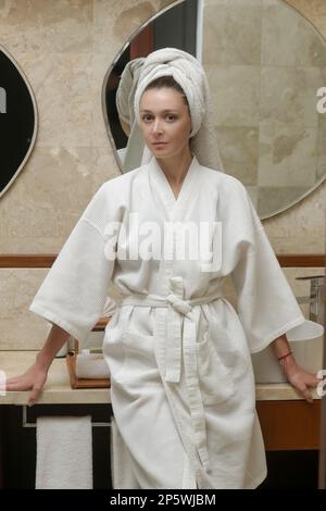 Ritratto di giovane donna in accappatoio di cotone bianco e capelli avvolti in asciugamano in bagno Foto Stock